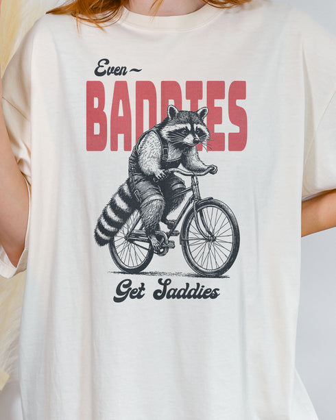 Close-up of woman wearing an ivory "Even Baddies Get Saddies" shirt with a funny retro graphic of a raccoon riding a bicycle.